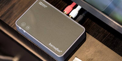 A close-up of the Bernafon TV-A TV adapter with power and pairing buttons, connected via red, white, and black cables, sits on a dark wooden surface next to a monitor.
